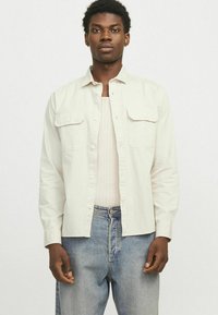 Camicia button-up beige chiaro con due tasche frontali, maniche lunghe e una vestibilità rilassata, indossata sopra una canotta a coste e abbinata a jeans blu.