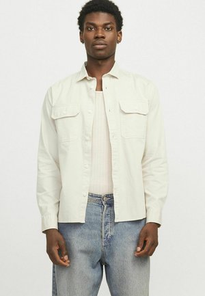 Camicia button-up beige chiaro con due tasche frontali, maniche lunghe e una vestibilità rilassata, indossata sopra una canotta a coste e abbinata a jeans blu.