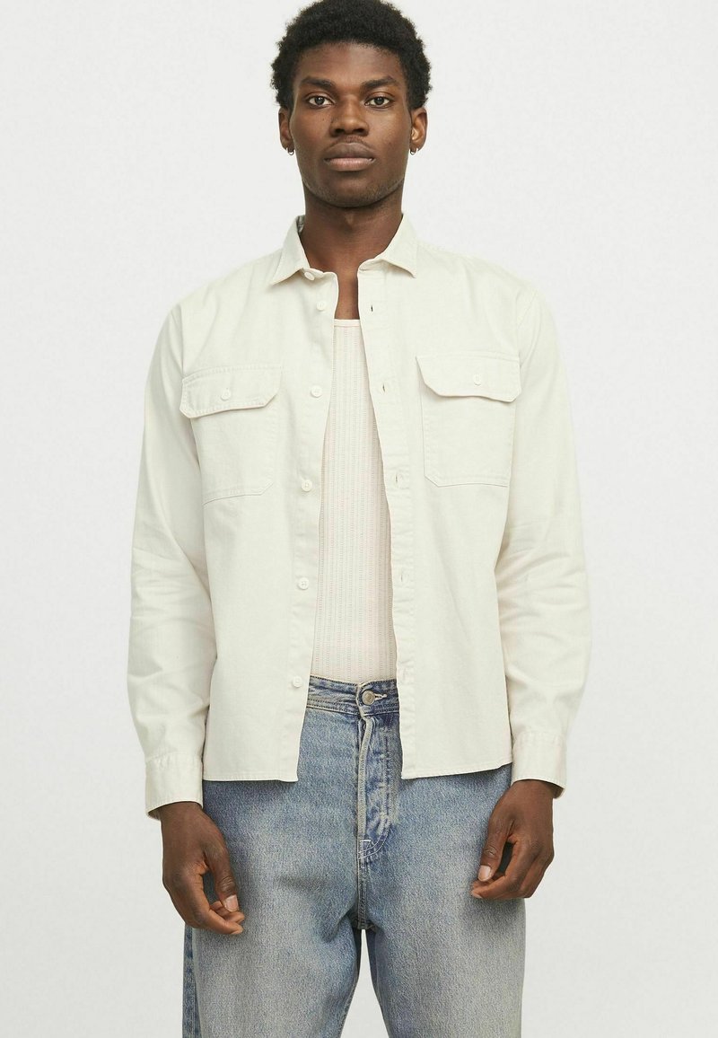 Camicia button-up beige chiaro con due tasche frontali, maniche lunghe e una vestibilità rilassata, indossata sopra una canotta a coste e abbinata a jeans blu.
