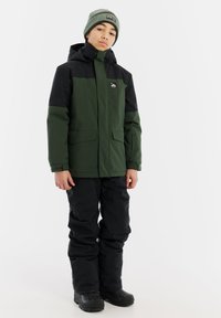 Grüne und schwarze isolierte Jacke mit Reißverschluss, zwei Seitentaschen und Kapuze, kombiniert mit schwarzen wasserdichten Hosen und schwarzen Stiefeln.