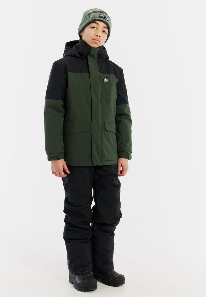 Grüne und schwarze isolierte Jacke mit Reißverschluss, zwei Seitentaschen und Kapuze, kombiniert mit schwarzen wasserdichten Hosen und schwarzen Stiefeln.