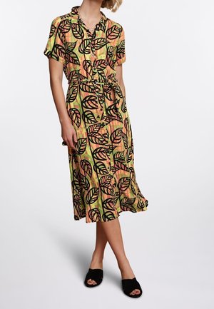 Femme portant une robe midi à manches courtes boutonnée avec des motifs de feuilles noires sur fond orange et vert, et des chaussures noires sans lacets.