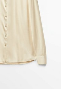 Chemise à manches longues beige clair à boutons, avec une texture lisse, présentant un ourlet droit et un léger éclat. Les boutons sont alignés verticalement.