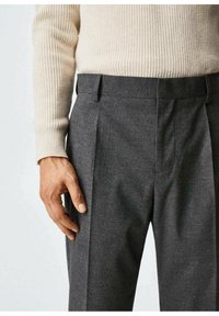 Main reposant sur un pantalon gris foncé sur mesure, associé à un pull en maille côtelée beige, sur un fond clair uni.