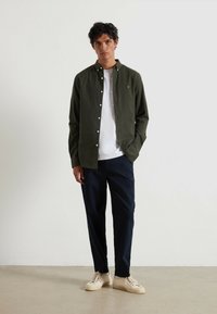 Ciemnozielona koszula z guzikami noszona na białej koszulce, w zestawie z granatowymi spodniami i kremowymi butami. Koszula ma małe logo oraz guziki.