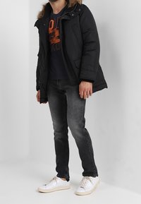 Svart vadderad jacka med foder i päls, bärs över en marinblå t-shirt med orange tryck, tillsammans med mörka jeans och vita sneakers.