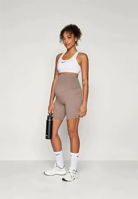 En fitte kvinne iført en hvit Nike sports-bh, høytlivede beige sykkelshorts, hvite Nike sokker og sneakers, holder en svart vannflaske.