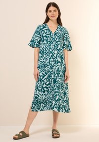 Teal-kuvioinen mekko, jossa on V-mallinen kaula-aukko, lyhyet volyymiset hihat ja napit edessä, yhdistettynä ruskeisiin sandaaleihin. Sileä kangas.