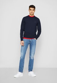 Un pull col rond bleu marine superposé à une chemise rouge, associé à un jean bleu clair et des baskets blanches, avec un petit logo sur le pull.
