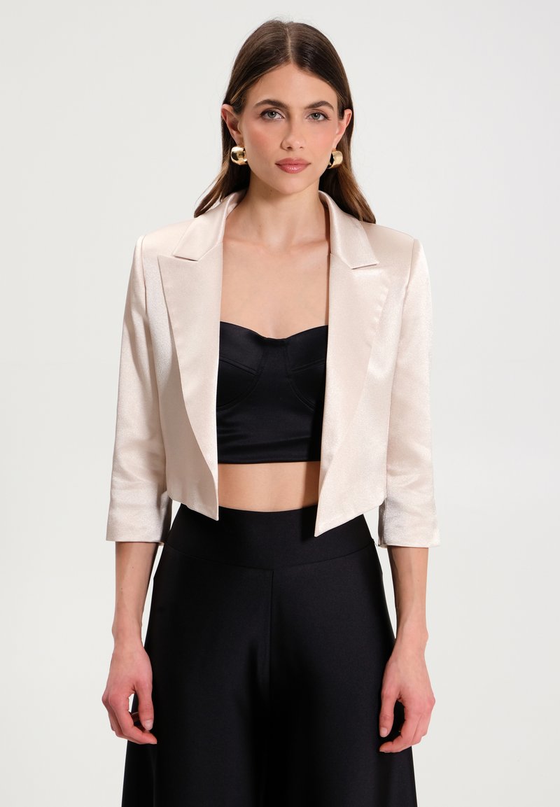Gekürzter Blush-Satin-Blazer mit Reverskragen und drei Viertelärmeln, kombiniert mit einem schwarzen, gekürzten Oberteil und hoch taillierten schwarzen Hosen.
