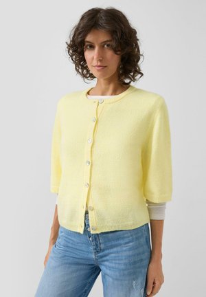 Vrouw met krullend haar, gekleed in een lichtgele korte mouw cardigan met knopen over een wit top met lange mouwen en lichtblauwe spijkerbroek.