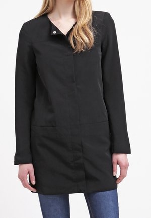 Femme portant un manteau noir sans col à manches longues avec un bouton-pression au niveau du cou, associé à un jean bleu.