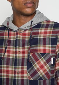 Červená a zelená kostkovaná flanelová košile s šedou mikinou. Obsahuje kapsu na hrudi se šikmým kostkovaným vzorem, zapínání na knoflíky a stahovací šňůrky.