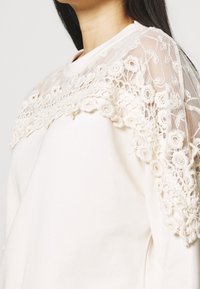 Crèmesweater met een doorschijnend kanten inzetstuk en bloemrijke borduurwerk die de halslijn accentueren. Lange mouwen en zacht katoenmateriaal.