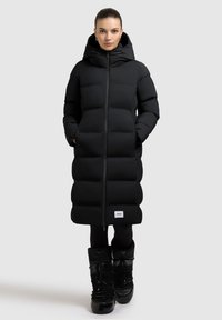 Schwarze Steppjacke mit Kapuze, gerader Schnitt, gestepptes Design und einem Logopatch am Saum. Kombiniert mit schwarzen Schneestiefeln.