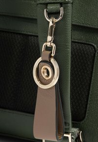 Detalle de un bolso de mano de cuero texturizado verde que incluye un clip de tono dorado, una etiqueta de cuero marrón y un adorno circular con marca.