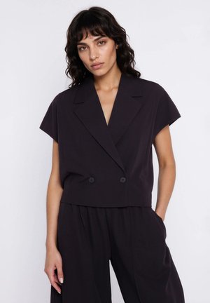 Kobieta o falowanych ciemnych włosach, ubrana w czarną marynarkę na krótki rękaw z podwójnymi guzikami oraz pasujące luźne czarne spodnie, stoi z jedną ręką w kieszeni.