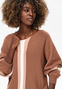 Vrouw met krullend haar met highlights, gekleed in een bruin-beige gestreepte trui en een gouden kruisvormige ketting, die bedachtzaam opzij kijkt.