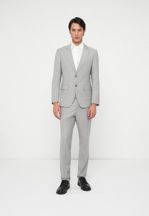 Šedý oblek s jedným gombíkom, klopovými reverzami a dvoma prednými vreckami, doplnený o bielu košeľu a čierne topánky.