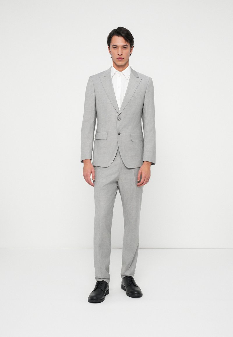 Fato cinza com fecho de um só botão, lapelas recortadas e dois bolsos frontais, combinado com uma camisa branca e sapatos pretos.