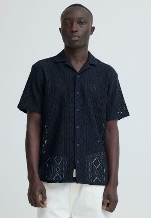 Man wearing a short-sleeve black button-up shirt with a diamond pattern, standing against a plain light background.