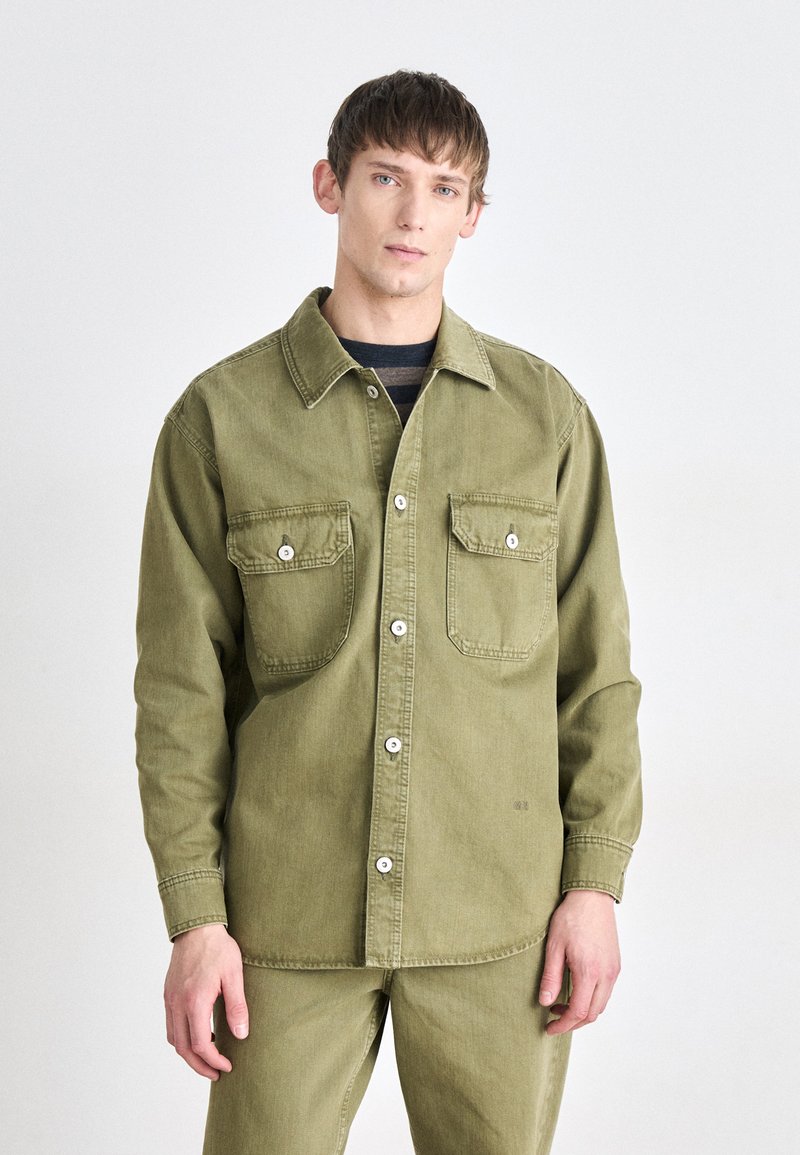 Chaqueta de camisa de algodón en verde oliva con dos bolsillos en el pecho y botones blancos. Presenta un cuello clásico, un ajuste relajado y mangas largas.