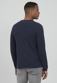 Pulover texturat de culoare bleumarin, cu maneci lungi, decolteu rotund si mansete ribbate, asociat cu pantaloni gri deschis. Vedere din spate.