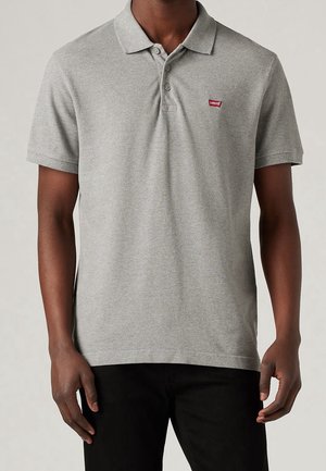 Mand iført en lysegrå kortærmet polo skjorte med et lille rødt Levi's logo på brystet og sorte bukser.