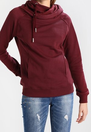 Nainen, joka pukeutunut viininpunaiseen huppariin, jossa on kiristysnyörit, sekä sinisiin kuluneisiin farkkuihin, joissa on silmukkakoristeet reiden kohdalla.