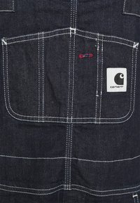 Dark denim overalls with white stitching, featuring a large back pocket. Carhartt logo patch on the lower right. Red accent stitching visible.