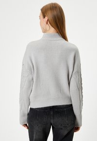 Pull en maille gris clair avec un col haut, des manches texturées présentant un design en torsades, et un ourlet côtelé, associé à un jean foncé.
