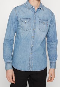 Ljusblå denimskjorta med traditionell krage, långa ärmar, två bröstfickor och guldknappdetaljer. Har en sydd fåll.