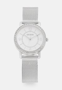 Montre à bracelet en maille argentée avec un cadran rond blanc, une lunette en cristal et des index en relief. Le nom de la marque "GUESS" est visible sur le cadran.
