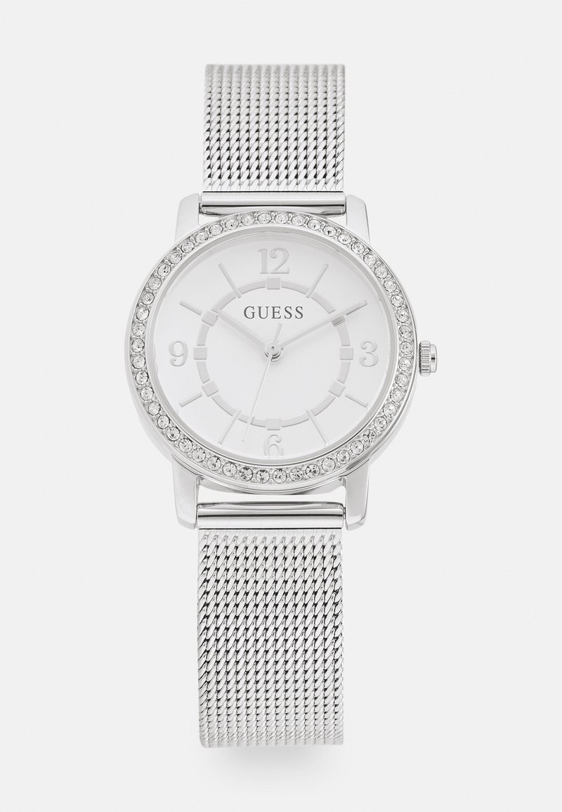 Montre à bracelet en maille argentée avec un cadran rond blanc, une lunette en cristal et des index en relief. Le nom de la marque "GUESS" est visible sur le cadran.