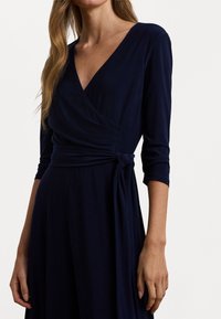 Abito avvolgente blu navy con scollatura a V, maniche a tre quarti e vita legata. Tessuto liscio con un design drappeggiato, che crea una silhouette fluida.