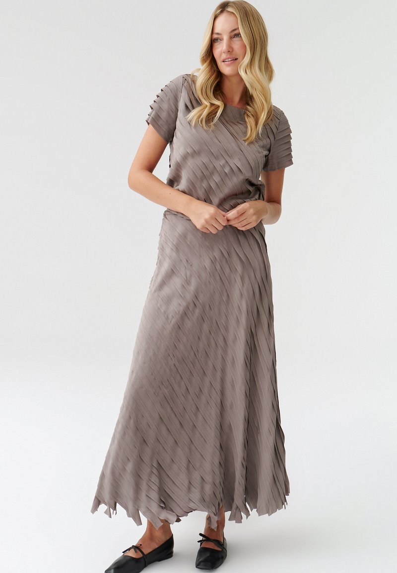 Pleated long dress in light taupe fabric, short sleeves, and uneven hem. Paired with black flat shoes, held at waist level.