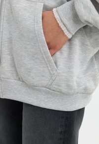 Sweatshirt gris en tissu texturé, avec une fermeture éclair et des poches latérales. Les poignets présentent un détail tricoté plus clair. Jeans sombres visibles en dessous.