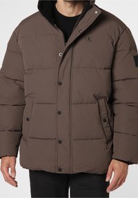 Brun puffjacka med dragkedja och knäppning, med två friska fickor, en logotyp på bröstet och en mjuk, texturerad yta.