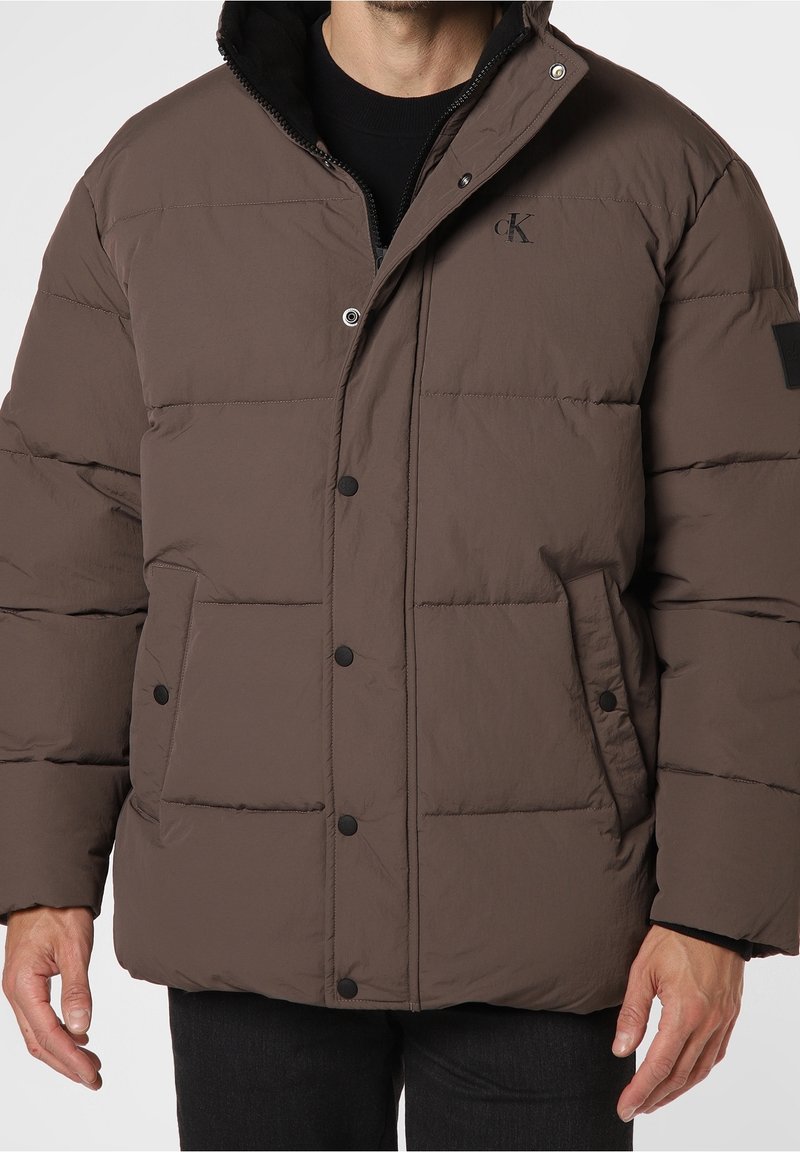 Brun puffjacka med dragkedja och knäppning, med två friska fickor, en logotyp på bröstet och en mjuk, texturerad yta.