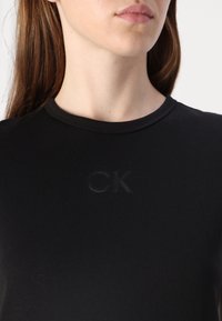 Fekete pamut póló kerek nyakkal, a mellrészen tone-on-tone "CK" logóval, amely domborúan van elhelyezve. Sima tapintású, testhez simuló fazon.