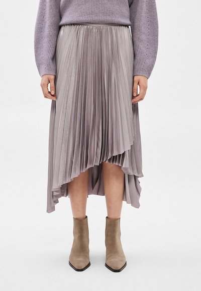 Femme portant une jupe midi plissée lavande, un pull en tricot violet et des bottines en daim beige, debout sur un fond blanc.