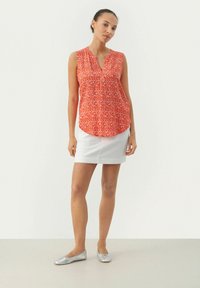 Orange ärmellose Bluse mit weißem abstraktem Muster, V-Ausschnitt, abgerundetem Saum; kombiniert mit einem weißen Rock und silbernen Ballerinas.