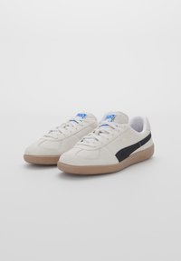 Sneaker in suede bianco con bottoni neri, punta arrotondata, chiusura con lacci e suola in gomma color gomma. Presenta un logo blu sulla linguetta.