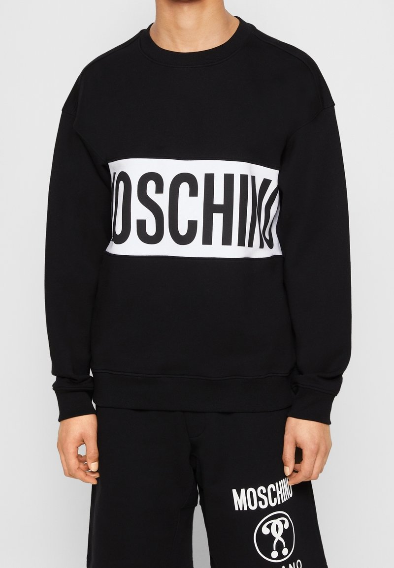 Felpa nera con una grande fascia bianca con la scritta "MOSCHINO" in grassetto nero. Polsini e orlo a coste, realizzata in tessuto morbido.