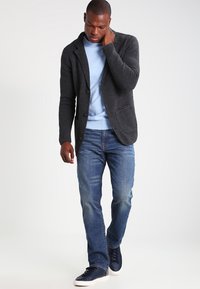 Mörkgrå texturerad cardigan över en ljusblå tröja, kombinerad med blå jeans och marinblå sneakers. Modellen står framför en neutral bakgrund.