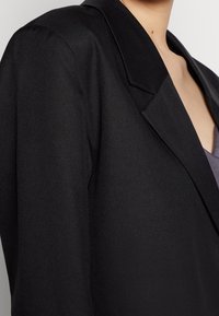 Svart blazer med strukturerade slag, textilerat tyg och skräddarsydd passform. Har en enkel knappstängning och minimalistiska designdetaljer.