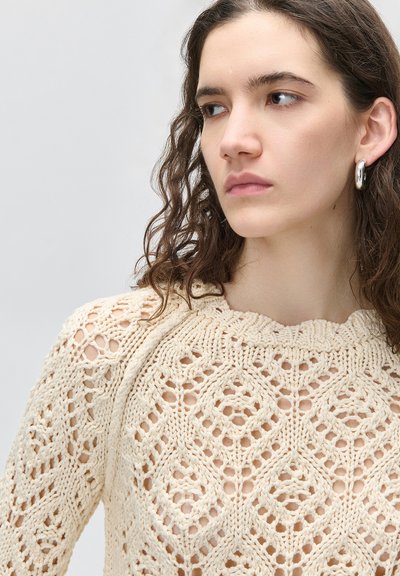 Νεαρή γυναίκα με κυματιστά καστανά μαλλιά, που φοράει ασημένια σκουλαρίκια κρίκους και μπεζ ανοιχτό πλεκτό πουλόβερ με σχέδιο, κοιτάζει προς τα πλάγια σε ουδέτερο φόντο.