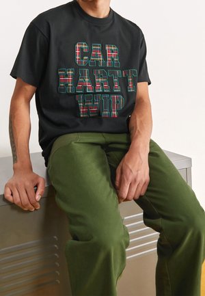 Mężczyzna siedzi na metalowej ławce, ubrany w czarną koszulkę z kraciastym napisem "CARHARTT WIP" oraz oliwkowe spodnie, ręce spoczywają na ławce i na nodze.