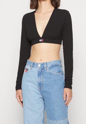 Femme portant un top court noir à manches longues avec un profond décolleté en V et un jean taille haute bicolore bleu clair et bleu moyen.