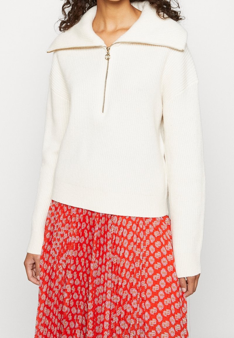 Cremefarbener, gerippter Pullover mit weitem Kragen und einem goldenen Reißverschluss vorne. Getragen über einem roten Rock mit einem weißen abstrakten Muster.